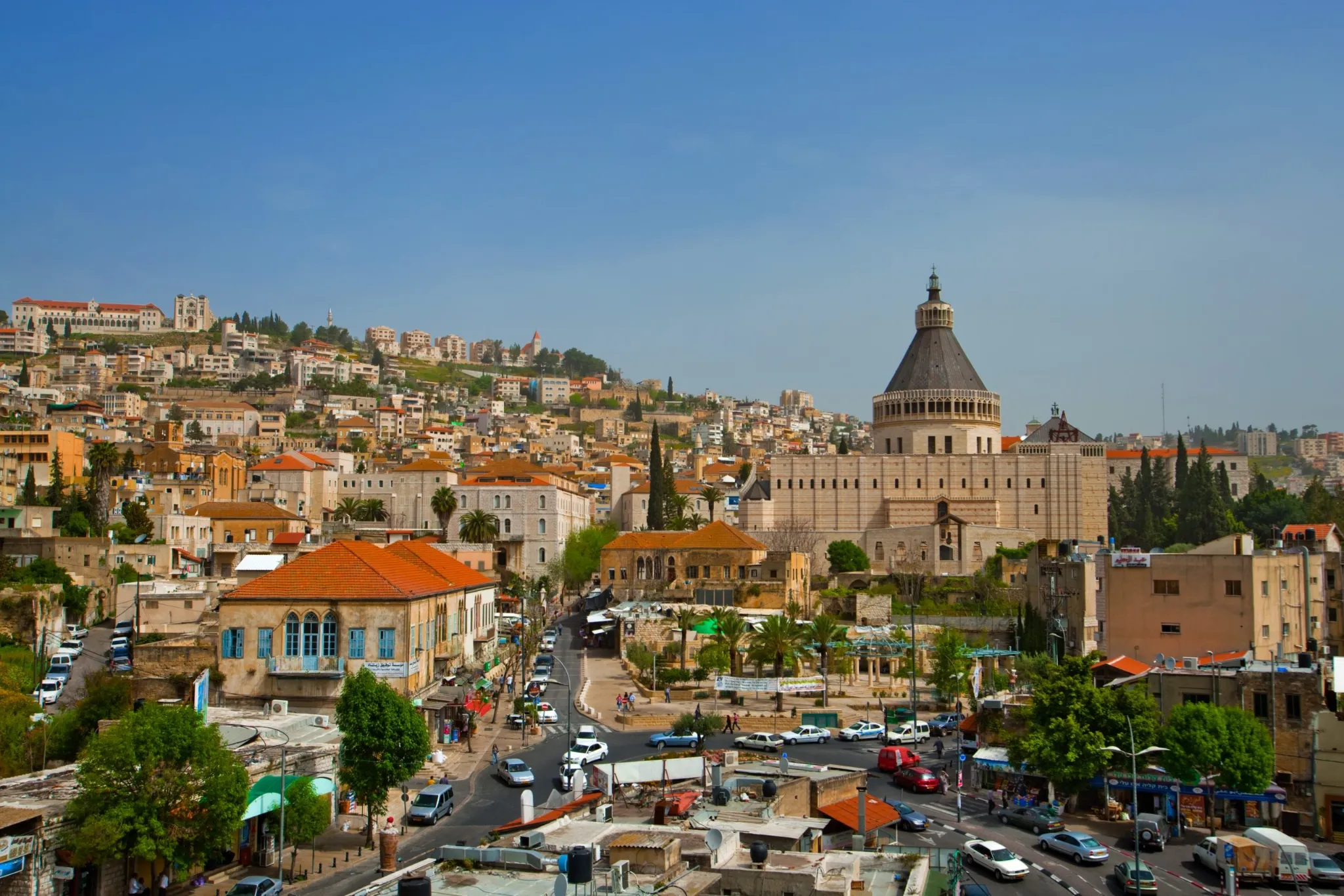 A cityscape view of Nazareth, Israel, dominated by the massive, dome-shaped Basilica of the Annunciation. The foreground features a busy town square and lower buildings with terracotta roofs, and the background shows residential homes climbing up the steep hillside.