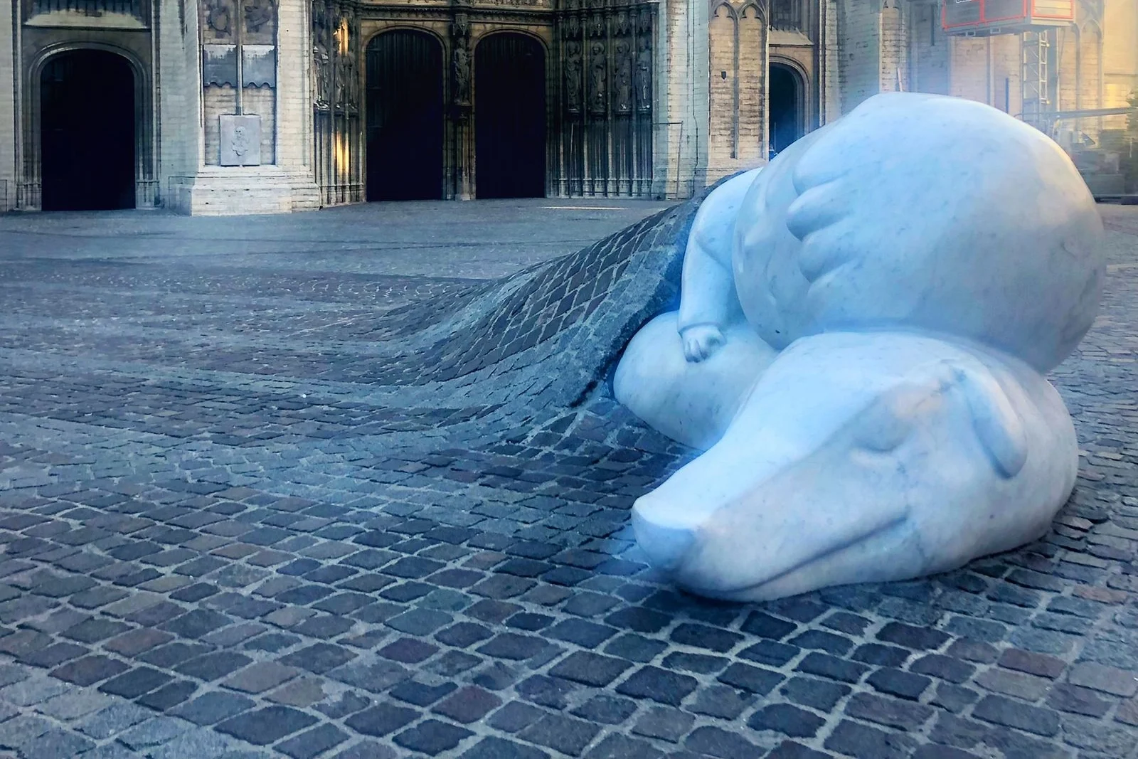 A contemporary white marble or stone sculpture of a sleeping, oversized polar bear on a cobblestone plaza. The bear appears to be emerging from or sinking into the ground, which ripples outward in cobblestones from beneath it. The setting is an open area near the dark, imposing stone entrance of a historic building.