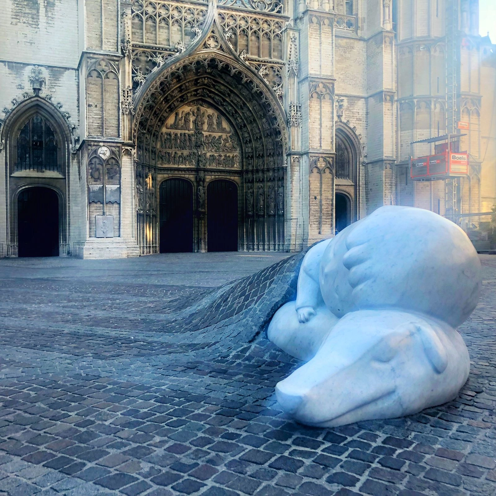 A contemporary white marble or stone sculpture of a sleeping, oversized polar bear on a cobblestone plaza. The bear appears to be emerging from or sinking into the ground, which ripples outward in cobblestones from beneath it. The setting is an open area near the dark, imposing stone entrance of a historic building.