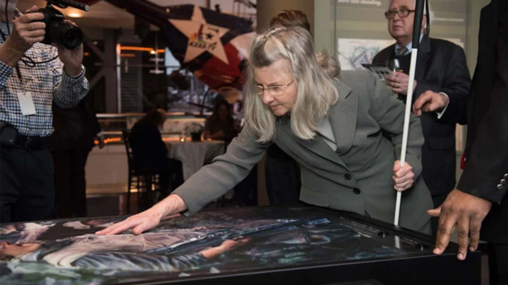 A middle-aged woman with glasses and blonde hair, wearing a gray suit, leans forward to gently run her hand across a large, tactile reproduction of a painting, likely in a museum setting. She is holding a white cane with her other hand, and in the background, other attendees and an aircraft propeller display are visible.