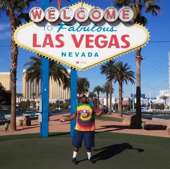 A person wearing a brightly colored tie-dye t-shirt and blue denim shorts stands directly in front of the iconic "Welcome to Fabulous Las Vegas Nevada" sign. The person has their arms spread wide and is smiling. The background shows palm trees and tall city buildings under a clear blue sky.