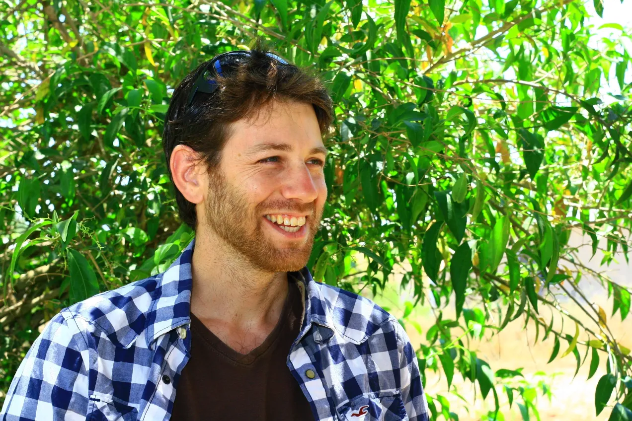 A close-up, sunlit portrait of a smiling young man with a short brown beard and sunglasses resting on his head. He is wearing a blue and white plaid, open shirt over a brown v-neck shirt. He is standing outdoors, framed by vibrant green leaves and foliage.