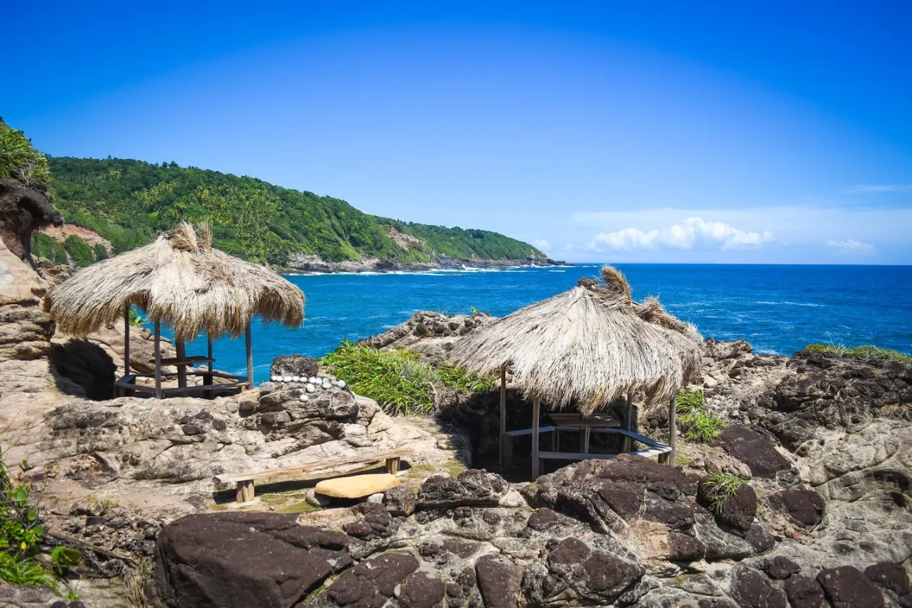 A scenic view of two small, rustic, open-sided gazebos with thatched roofs, situated on a dramatic, rocky outcrop overlooking the bright blue ocean. In the foreground, dark, rough boulders and a small wooden bench are visible. The background shows the rugged coastline covered in green vegetation under a clear, deep blue sky.