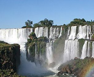 A multi-teared waterfall cascades churning white water, among rocks and trees, under a blue sky.
