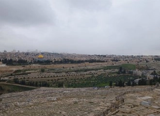 A panoramic view over the Kidron Valley and the Old City of Jerusalem under an overcast sky. In the foreground is the ancient Jewish Cemetery on the Mount of Olives. The middle ground features the Garden of Gethsemane, and the background shows the skyline of Jerusalem, dominated by the yellow-gold Dome of the Rock.