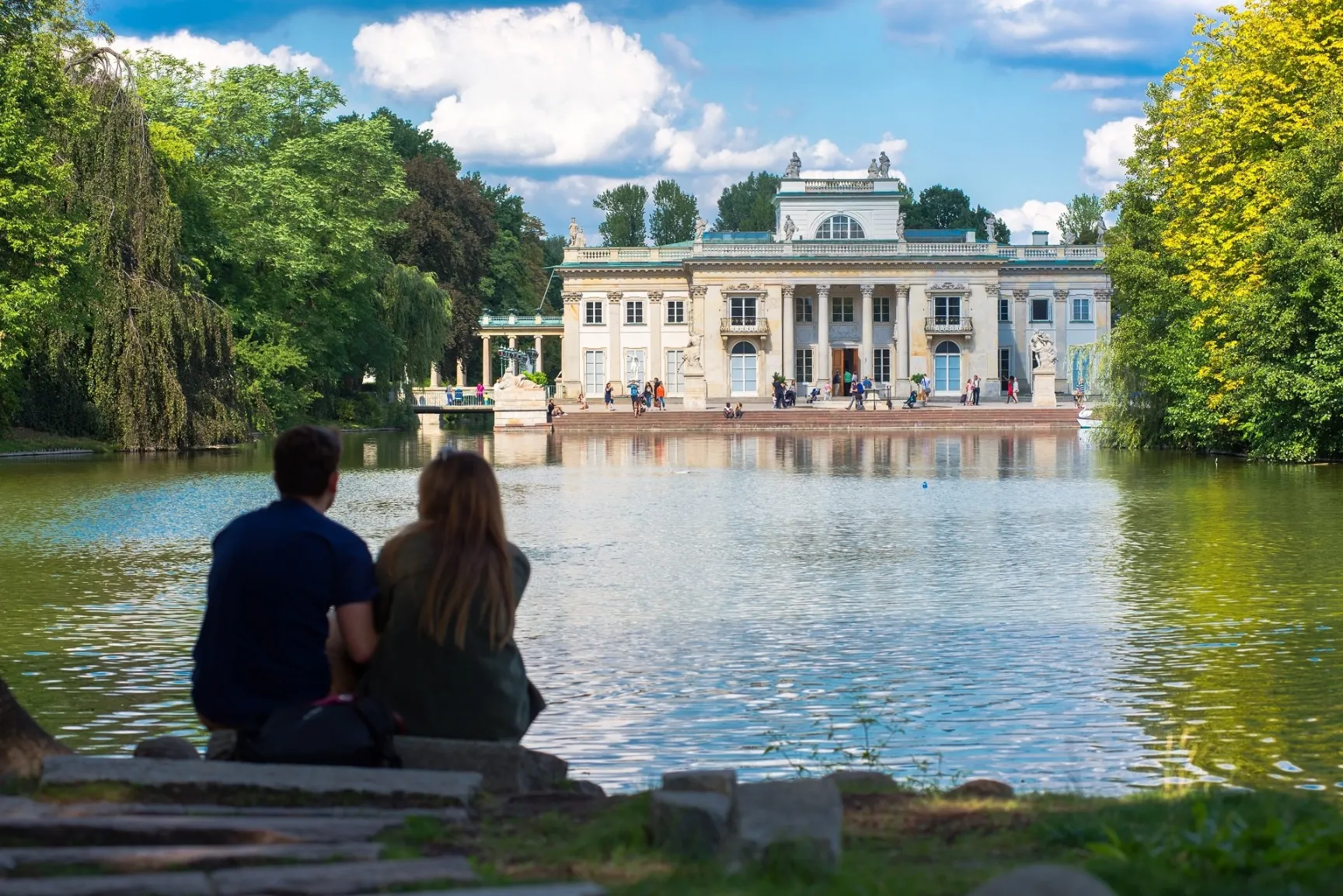 A serene view across a large pond in a park, showing the Palace on the Isle in Warsaw, Poland. In the foreground, a couple sits on a stone ledge with their backs to the camera, looking toward the water. The grand, white neoclassical palace is perfectly reflected in the calm lake, framed by dense green trees and a bright blue sky with fluffy white clouds. Several people are visible strolling along the palace terrace in the distance.