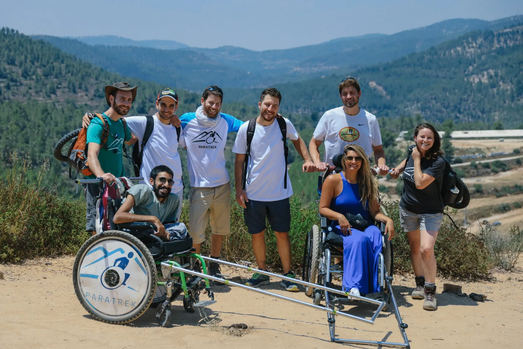 A group of eight happy people poses for a photo on a sunny mountain trail overlooking a vast forested valley. Two people are seated in specialized all-terrain wheelchairs, one man on the left whose chair displays the 'PARATREK' logo, and one woman on the right in a long blue dress. They are surrounded by six standing people, most wearing white t-shirts, suggesting they are a hiking support group.