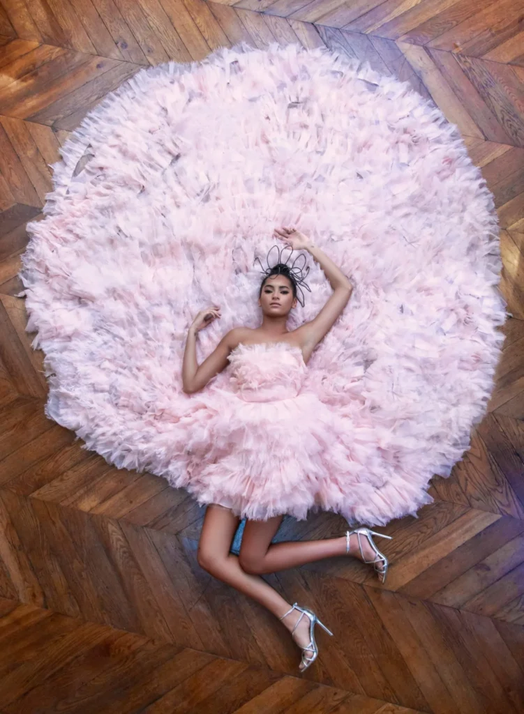An overhead photo of a woman wearing a voluminous, strapless, light pink ruffled tulle dress, lying on a herringbone wood floor. The skirt fans out completely around her in a large, fluffy circle. She is wearing silver high heels and a small decorative black headpiece.