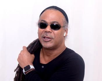 A close-up portrait of a man with dreadlocks, wearing sunglasses, a black beanie, and a black shirt with a buttoned collar. He is holding his dreadlocks over his shoulder with one hand, which wears a smartwatch. He has a subtle smile and appears to be speaking.