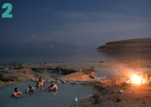 A panoramic, night-time shot of a social gathering on a beach next to a body of water. A bonfire burns brightly in the sand, illuminating a small group of people sitting around it. In the background, the dark, massive outline of mountains rises against a starry or dark-skied horizon. This is likely a scene at the Dead Sea or near one of the desert hot springs.