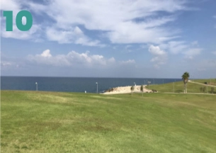 A horizontal landscape view showing a wide, flat expanse of green lawn sloping slightly toward the sea. The blue sea meets the horizon under a partly cloudy sky, and a small paved area with lamp posts is visible near the water.