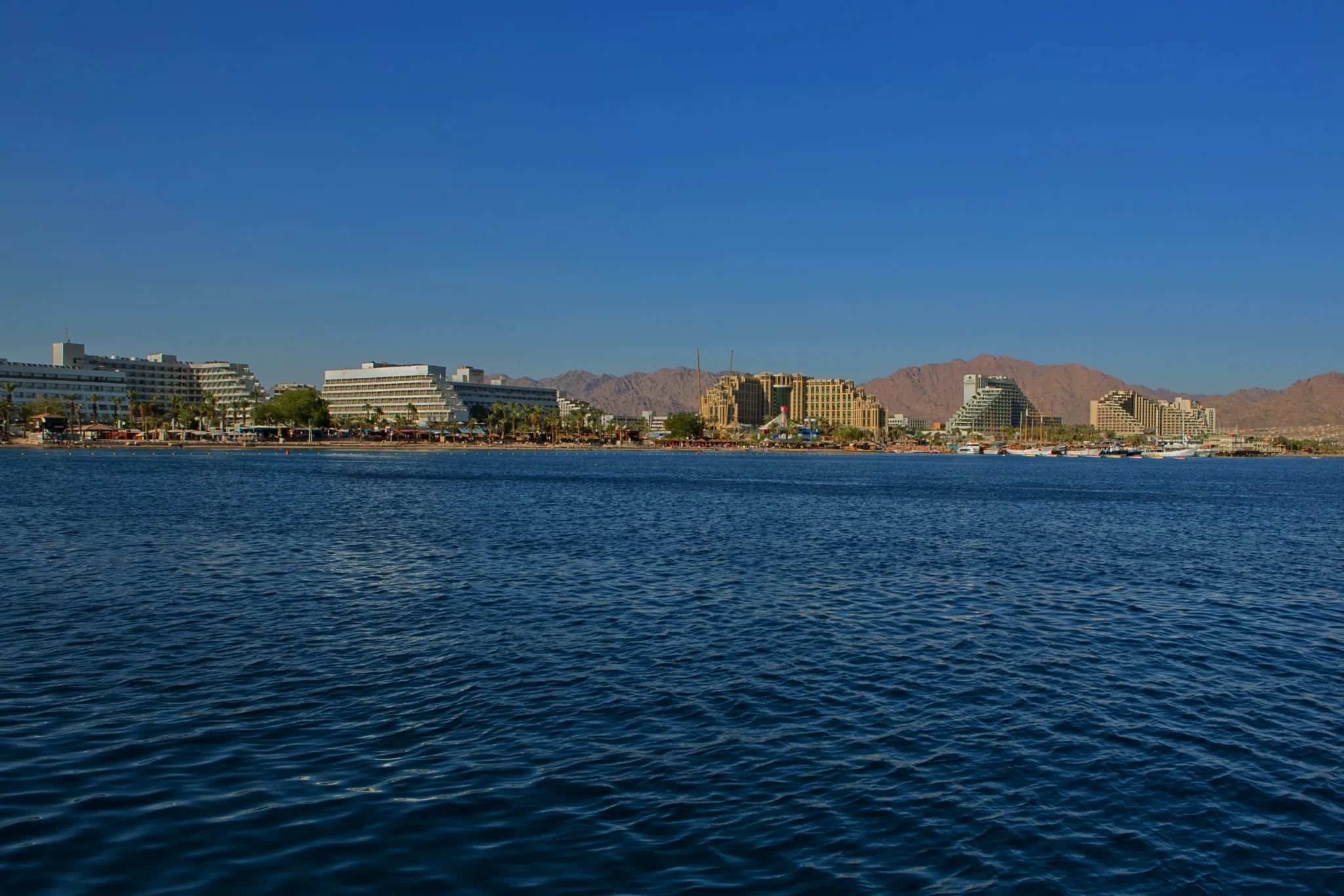A panoramic view of the coastline of a resort city, likely Eilat, Israel, showing several large, modern hotel buildings lining the shore. The deep blue sea is in the foreground, and the background features rugged, dry brown mountains against a clear blue sky.