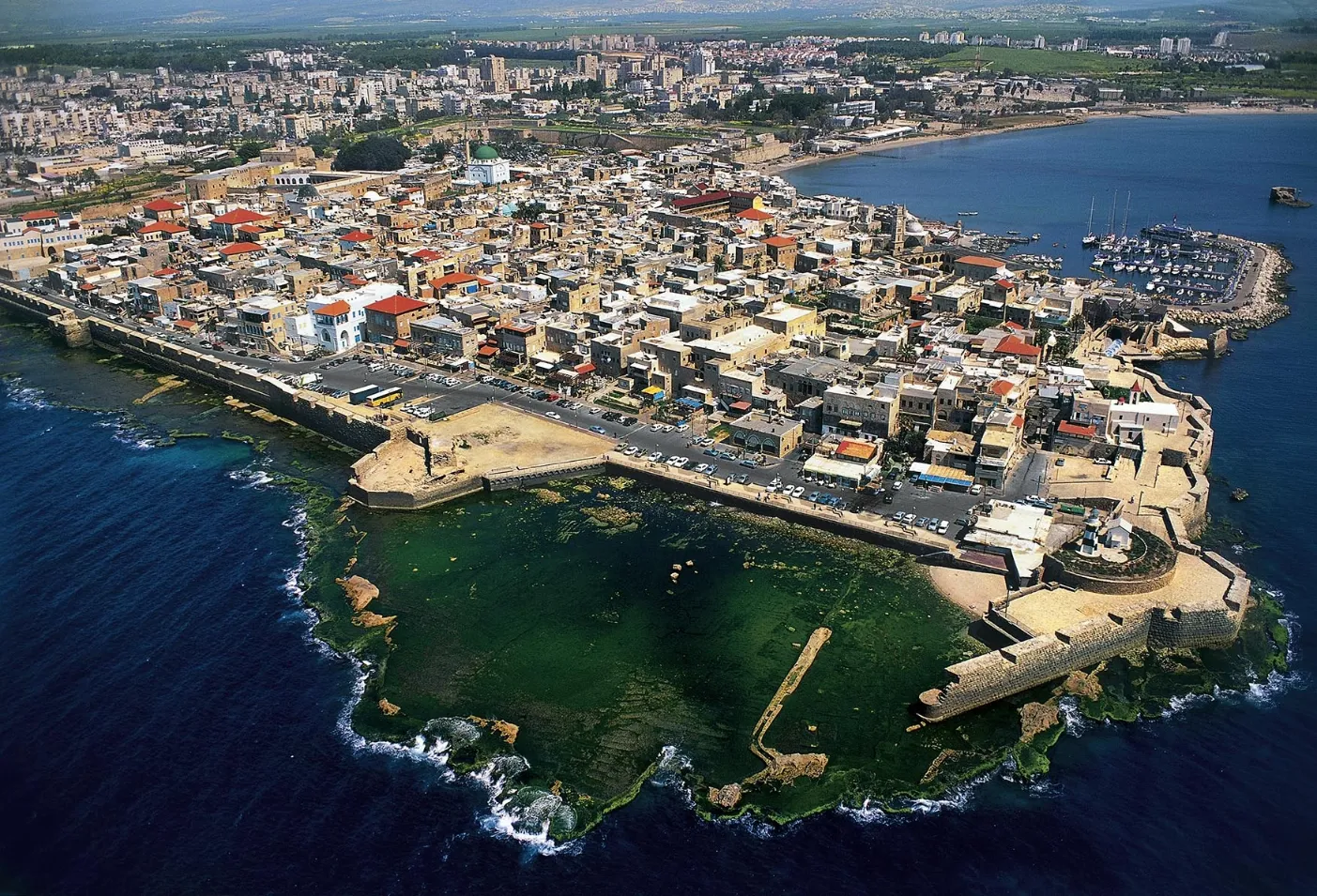An aerial, wide-angle view of a densely built, ancient coastal city on a peninsula jutting into the dark blue sea, likely Acre (Akko), Israel. The city is surrounded by heavy stone fortifications and features numerous low buildings with terracotta roofs and a prominent mosque dome. A modern city extends along the shoreline in the background.