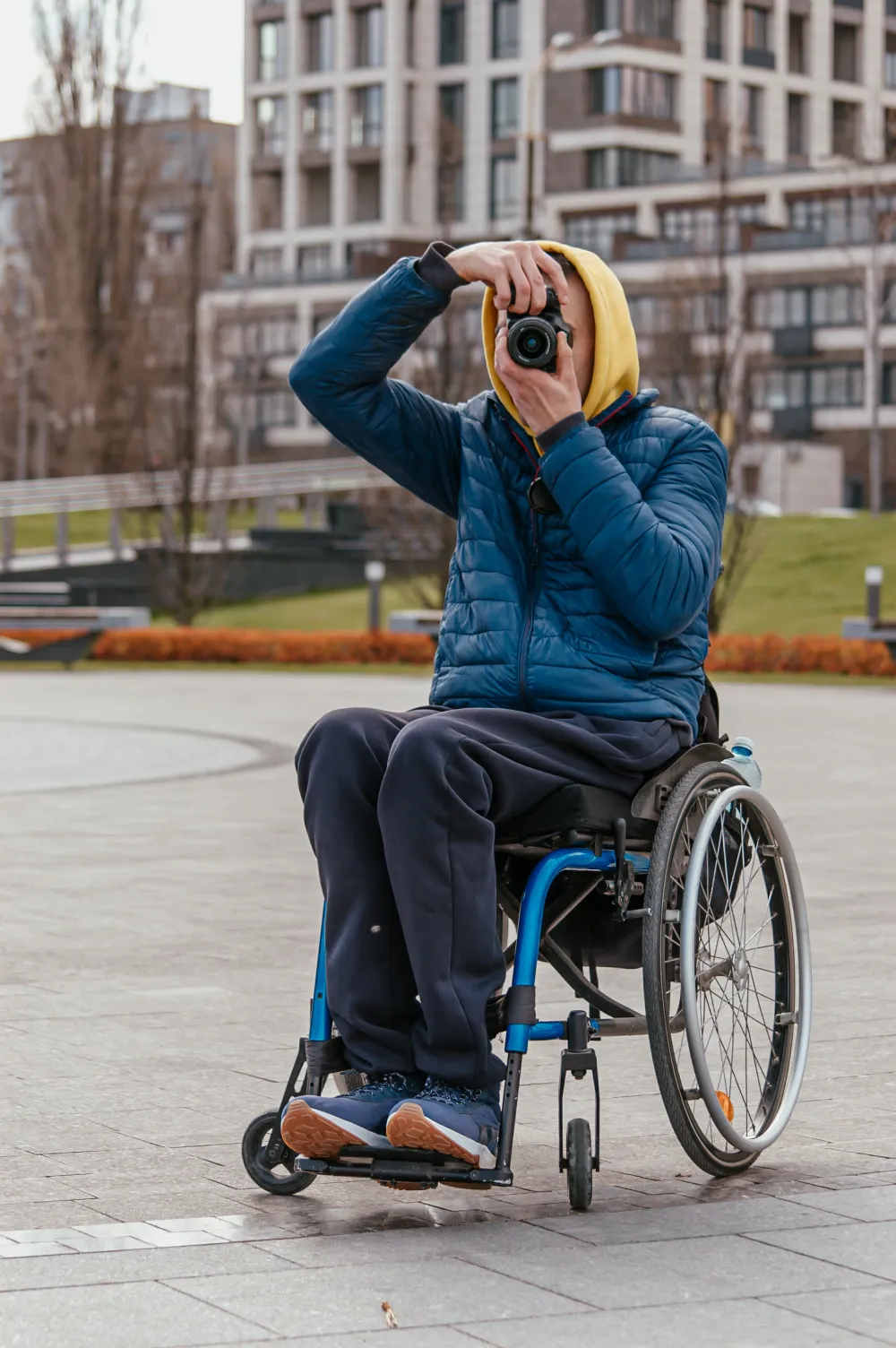 A person sitting in a modern, blue-framed wheelchair is actively taking a photo with a black DSLR camera, holding it up to their eye. They are wearing a blue puffer jacket, dark pants, and have a yellow hood pulled up. They are outdoors in an urban setting on a paved plaza, with modern apartment buildings visible in the blurred background.