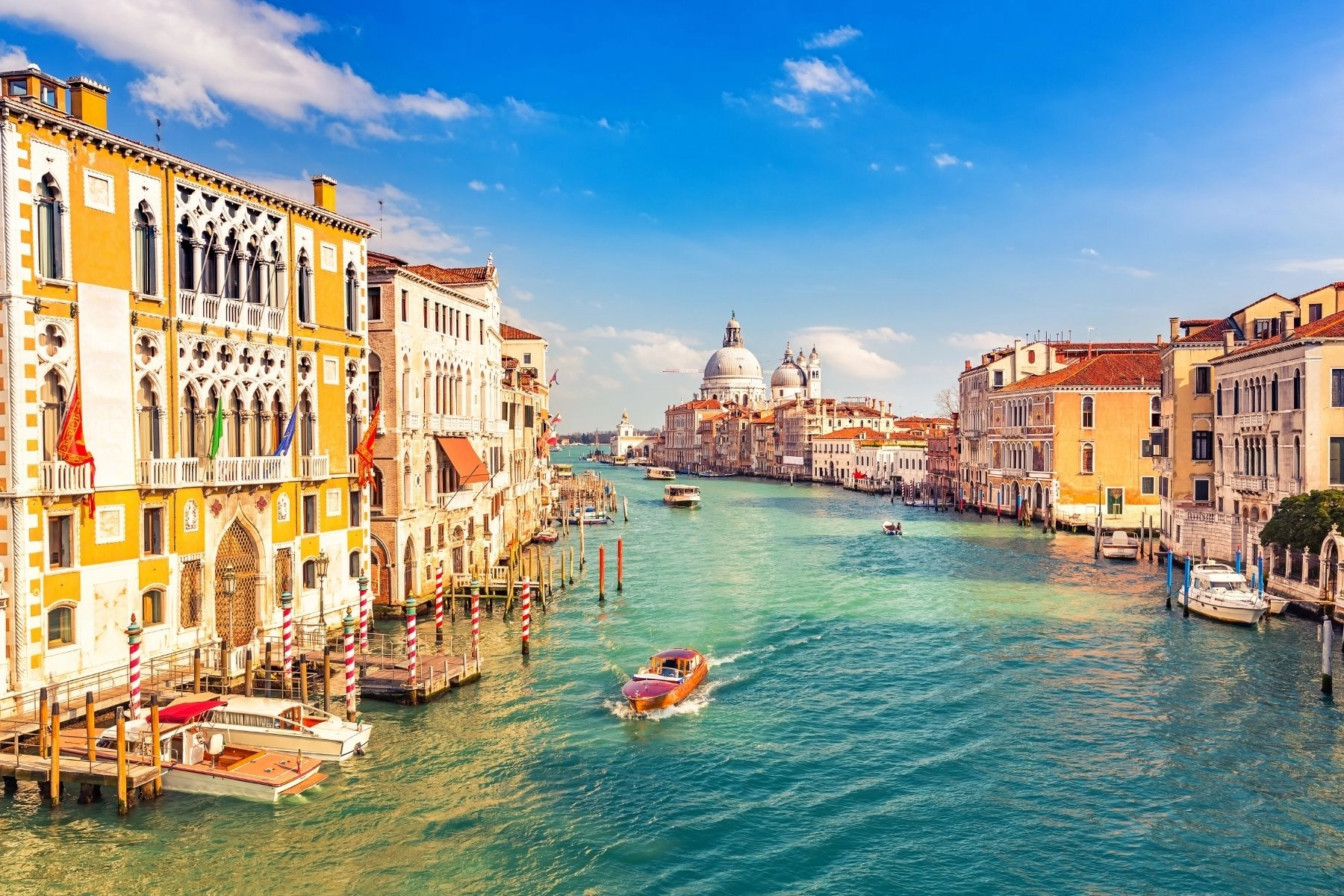 A vibrant, sunny photo of the Grand Canal in Venice, Italy. The turquoise water is filled with several boats and water taxis, one of which is speeding toward the viewer. The canal is lined with colorful, historic Venetian palaces and buildings in shades of white and ochre. In the distance, the iconic domed church of Santa Maria della Salute is prominently visible under a bright blue sky with white clouds.