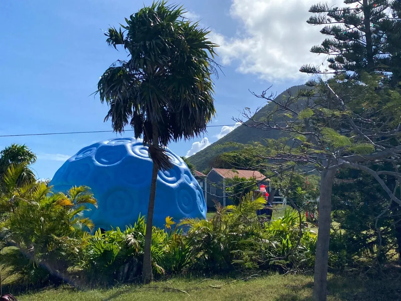 A bright blue, dome-shaped structure with raised swirling patterns, resembling a giant orb or planet model, is nestled among lush green tropical foliage and palm trees. In the background, a small, gray wooden building is partially visible, with a large, verdant, conical mountain rising against a blue sky with white clouds.