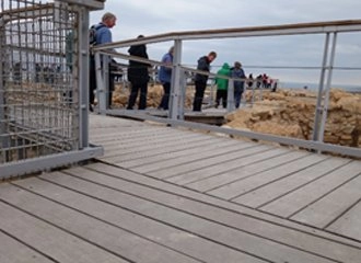 A wooden boardwalk with metal railings and scaffolding, built over archaeological ruins at the top of Masada. Several tourists are standing along the railing, looking out over the landscape and the exposed stone ruins.