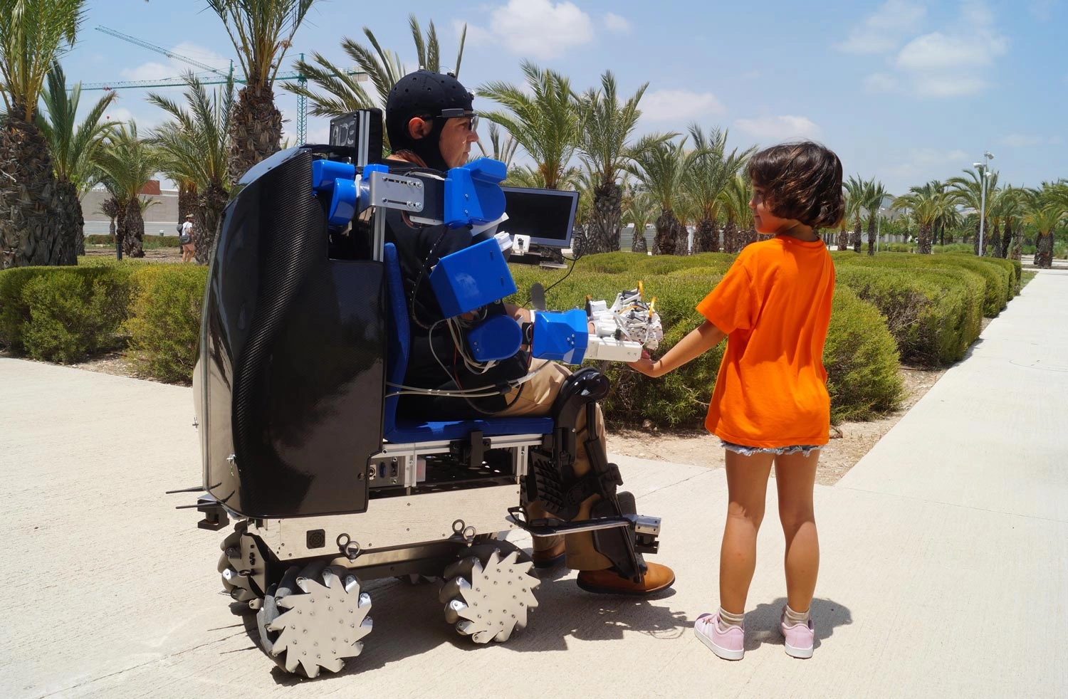A man wearing a black helmet and seated in a sophisticated, motorized, off-road wheelchair with a black carbon fiber shell and blue robotic arm attachments is interacting with a young girl. The man is looking at the girl, who is wearing a bright orange t-shirt and shorts. They are outdoors on a sunny day on a paved path, surrounded by palm trees and manicured green hedges. The chair features large, unusual, star-shaped wheels.