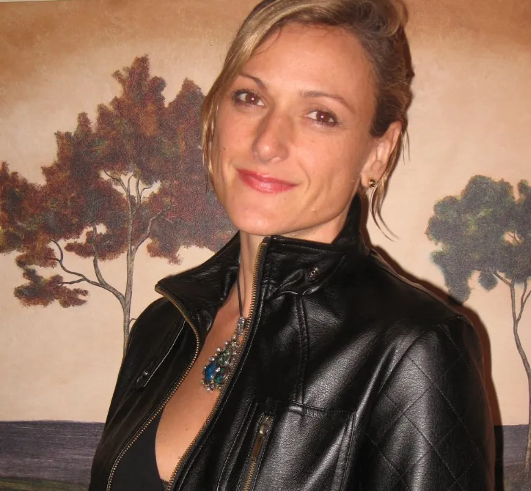 A headshot of a woman with blonde hair pulled back, smiling slightly at the camera. She is wearing a black leather jacket with the zipper partially open, revealing a large, ornate blue and green pendant necklace. A painted mural featuring stylized brown and dark green trees on a pale background is visible behind her.