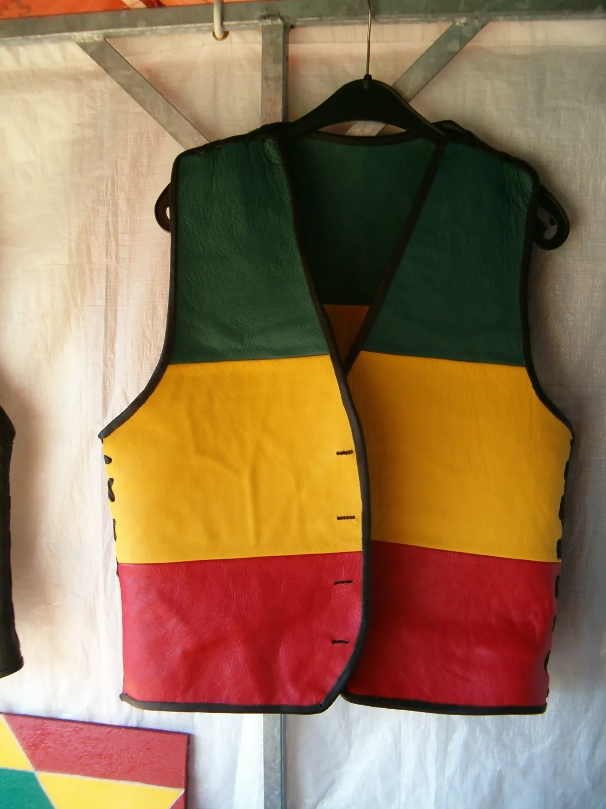 A sleeveless vest made of leather or fabric, hanging from a rack. The vest is horizontally color-blocked in the Rastafarian colors: green on the top, yellow in the middle, and red on the bottom. The edges are trimmed in black.