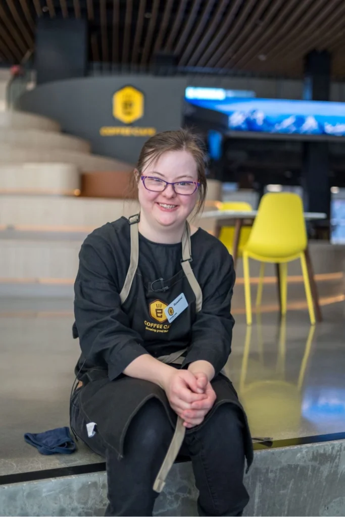 A portrait of a young woman with Down syndrome, wearing glasses and a black shirt with a black and tan "Coffee Cafe" apron, smiling brightly at the camera while sitting indoors. The background shows a multi-level coffee shop space with a yellow chair and wooden steps. Her name tag reads "Rachael."