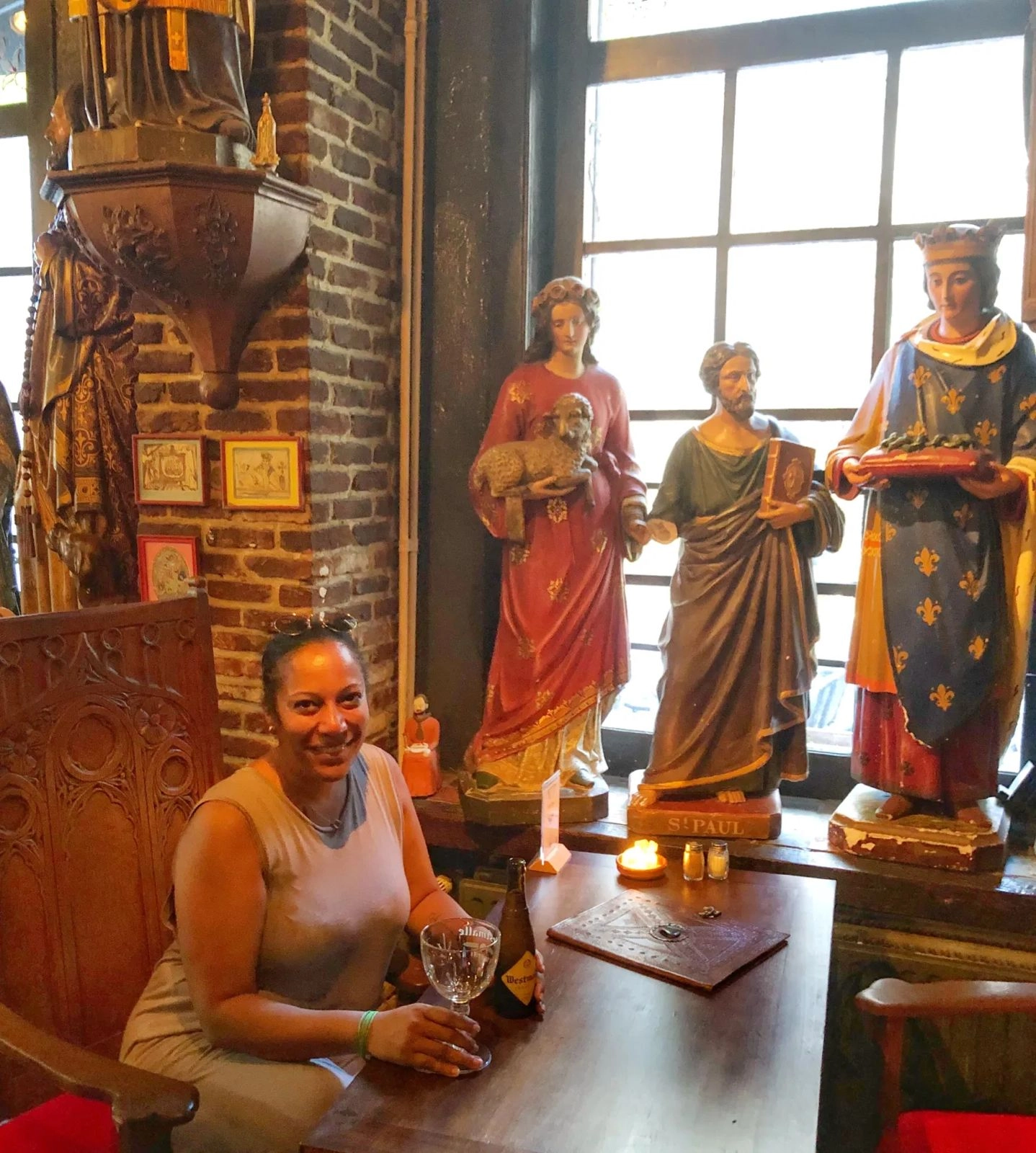 A woman is sitting and smiling at a wooden table in a dimly lit, rustic interior, possibly a historic pub or cafe. She is holding a stemmed glass next to a beer bottle. Behind her, against a large window, stand three tall, painted wooden statues of religious figures: one holding a lamb, one labeled "St. Paul" holding a book, and one wearing a crown and holding a tray. The walls are exposed brick.