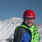 A close-up shot of a person smiling, wearing a red helmet and a bright neon green vest over a blue jacket, standing in front of a vast, snow-covered mountain range under a clear blue sky.