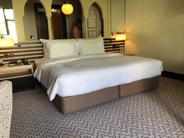 An opulent hotel bedroom featuring a large king-size bed with white linens and a tan base, set against a dark-striped headboard. The room is decorated in a rich, Middle Eastern style with arched doorways, a hanging lantern, and a patterned carpet.