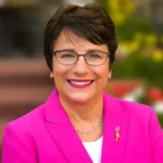 A headshot of a smiling woman with short, dark hair and glasses, wearing a vibrant fuchsia or bright pink blazer over a white shirt. She has a subtle pink ribbon or pin on her lapel and a gold necklace. The background is blurred, showing soft colors and architectural elements, suggesting an outdoor or professional setting.