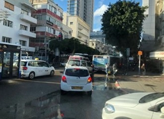 A street-level view of a busy intersection in an urban area, likely Tel Aviv, with wet pavement reflecting the light. There are multiple cars and buses stopped in traffic, including a white taxi and a white hatchback. Tall, modern and older apartment buildings line the street.