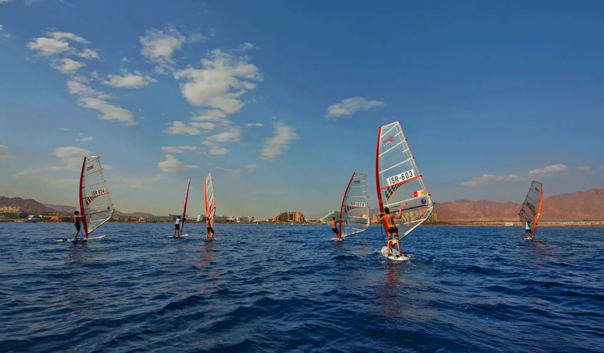 A wide-angle shot showing several windsurfers on the dark blue water, carrying red and white sails. The windsurfers are spread out across the sea, with a coastal resort town and a chain of barren mountains visible in the distance under a blue sky with scattered clouds.