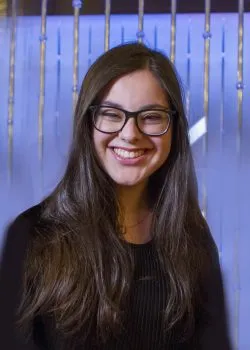 A portrait of a young woman with long, dark hair and glasses, wearing a black top. She is smiling broadly at the camera and is positioned in front of a blurred background with vertical blue and gold accents.