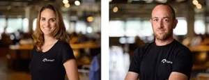 A side-by-side photograph of a woman and a man, both wearing black t-shirts with a white logo on the chest. The woman, on the left, has long brown hair and is smiling, while the man, on the right, is bald and bearded, also looking forward with a slight smile. The background appears to be a blurred indoor office or shared workspace.