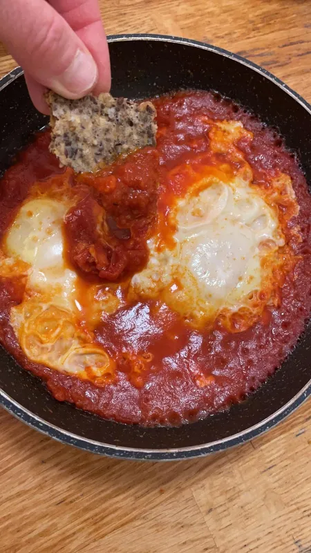 A close-up, overhead shot of a black pan containing shakshuka, a dish of two eggs poached in a bright red tomato sauce. A hand is dipping a piece of dark, seeded bread into the sauce and egg mixture.