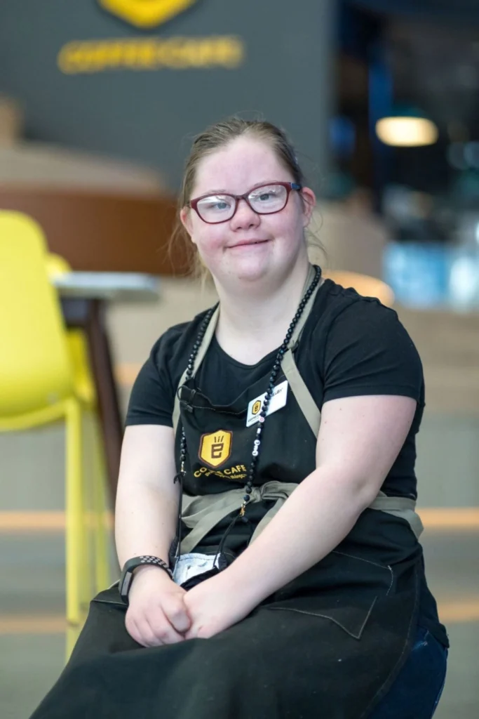 A portrait of a young woman with Down syndrome, wearing glasses and a black shirt with a black and tan "Coffee Cafe" apron, smiling brightly at the camera while sitting indoors. The background shows a multi-level coffee shop space with a yellow chair and wooden steps. Her name tag reads "Rachael."