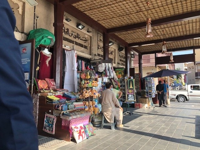 The shaded entrance to a traditional market or souk shop, displaying various items like colorful textiles, small stuffed toys, and souvenirs. The shop has traditional Arabic script above the entrance and a straw mat ceiling. A man in traditional clothing is sitting on a low stool near the display.