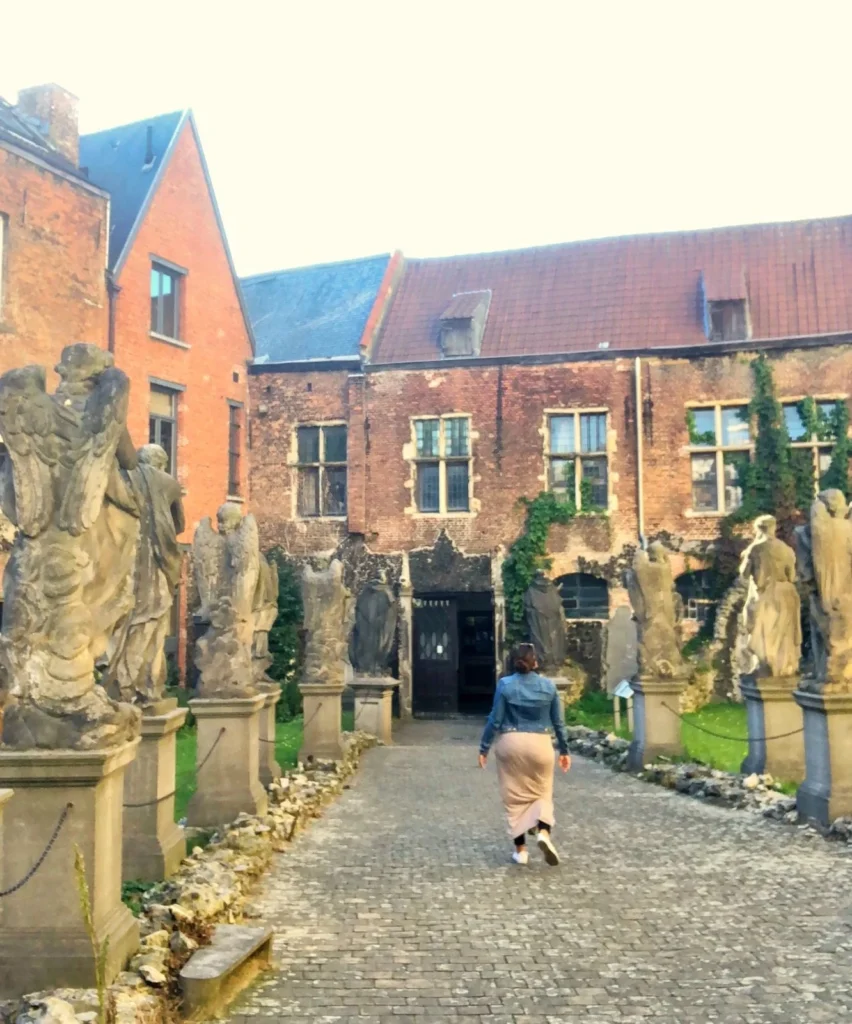 A vertical shot of an outdoor courtyard or garden with a cobblestone path lined by rows of weathered stone statues of angels or religious figures on pedestals. A woman wearing a denim jacket and a light skirt is walking away down the path towards a doorway in a cluster of historical brick buildings with tiled roofs under a bright sky.