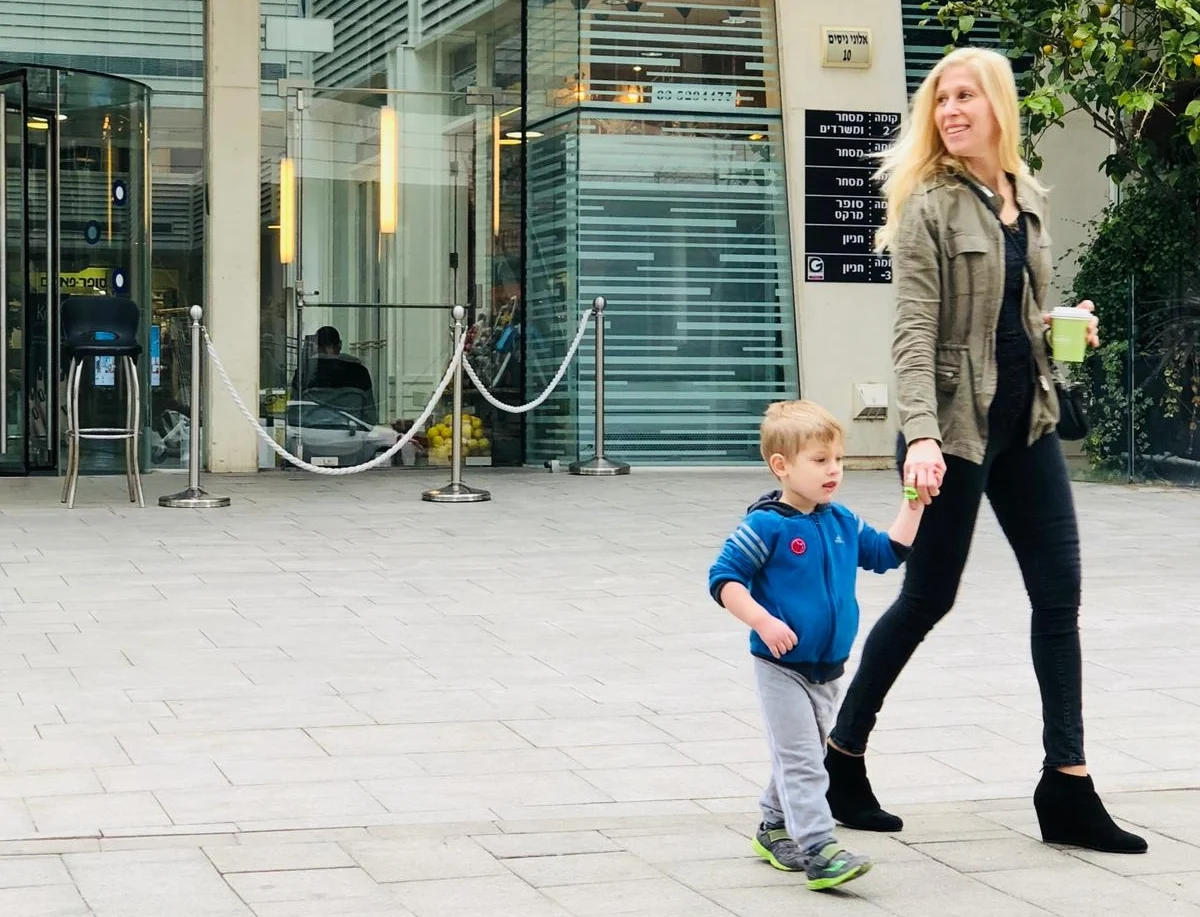 A blonde woman in a khaki jacket and black pants holds the hand of a small blonde boy in a blue zip-up jacket and gray pants as they walk across a stone sidewalk in front of a modern glass office building. The woman is smiling and holding a green cup in her other hand.