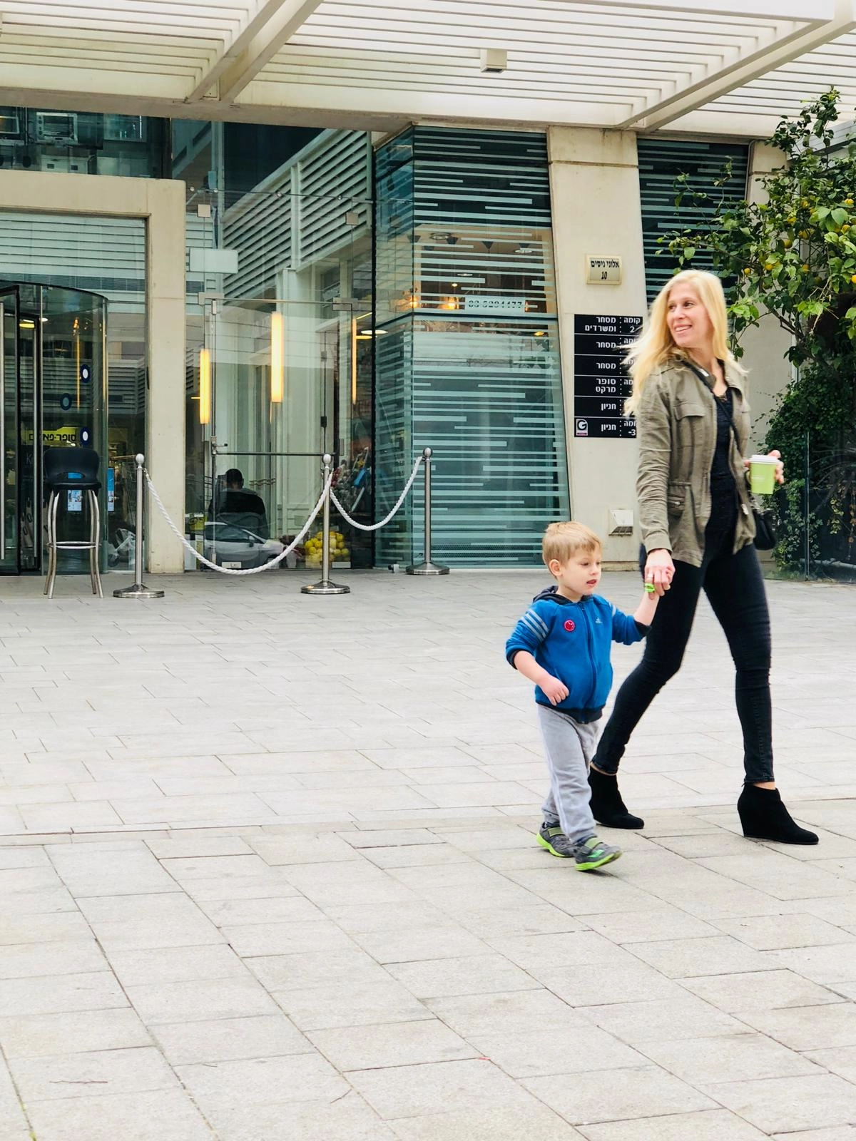 A blonde woman in a khaki jacket and black pants holds the hand of a small blonde boy in a blue zip-up jacket and gray pants as they walk across a stone sidewalk in front of a modern glass office building. The woman is smiling and holding a green cup in her other hand.