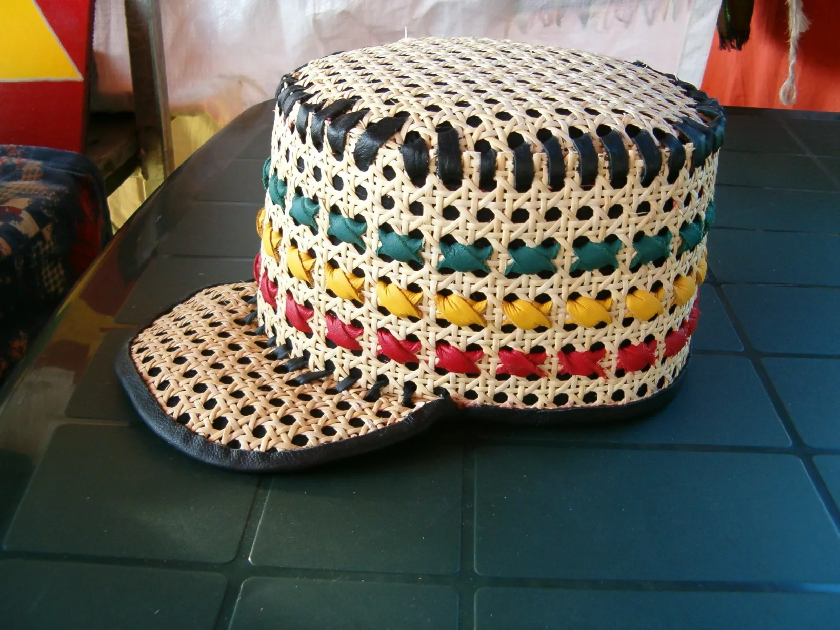 A handcrafted woven cane hat with a flat top and a short brim, sitting on a dark green table. The hat is decorated with woven strips of leather or fabric in black, red, yellow, and green, following a pattern of the Rastafarian flag colors. The brim is trimmed with black leather.