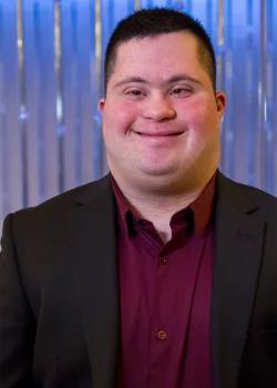 A close-up portrait of a man with Down syndrome wearing a dark blazer over a burgundy dress shirt, smiling directly at the camera. He is standing in front of a blurred, shimmering blue and white background.