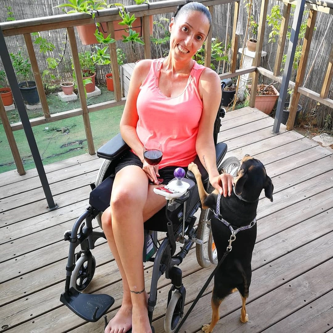A woman with dark hair pulled back, wearing a pink tank top and black shorts, sits barefoot in her motorized wheelchair on a wooden deck. She is smiling at the camera and holding a glass of red wine in one hand, while stroking a small black and tan dog with the other. The dog, wearing a leash, is standing on its hind legs with its paws resting on the armrest of the wheelchair. The background features a wooden railing covered with potted plants and greenery.