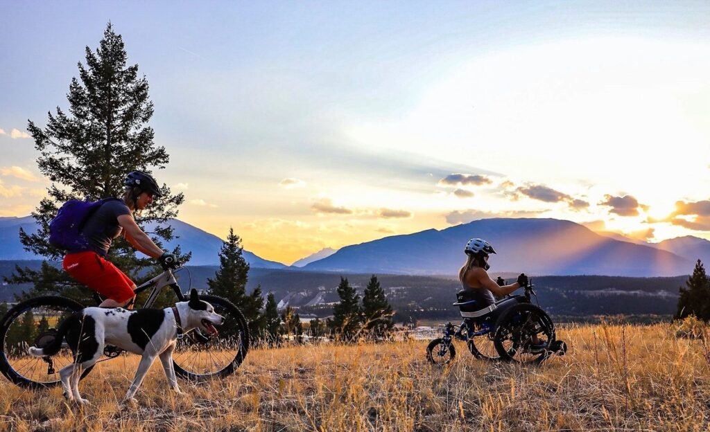 The sun sets over a distant mountain range. In the foreground, a person on a bicycle and dog follow a person in a 3-wheeled chair over the scrub.