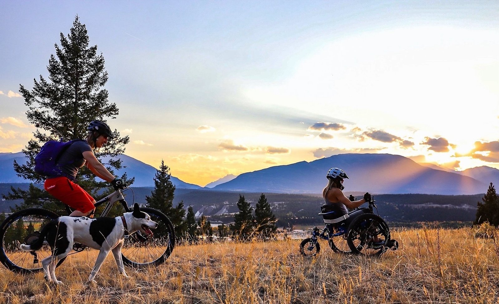 The sun sets over a distant mountain range. In the foreground, a person on a bicycle and dog follow a person in a 3-wheeled chair over the scrub.