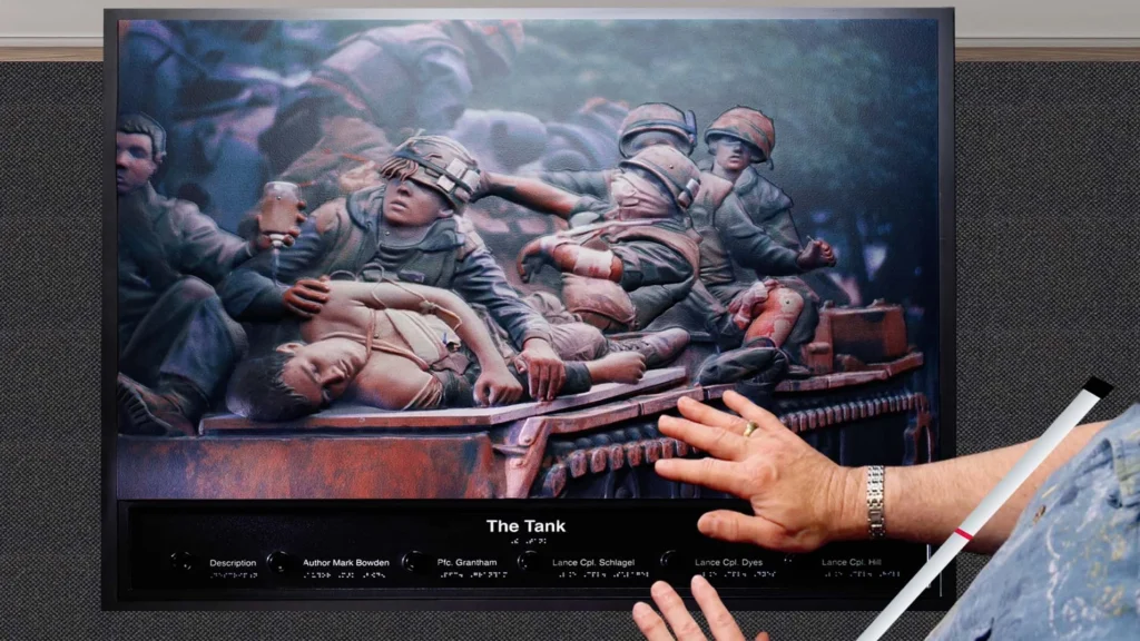 A close-up of a high-relief, three-dimensional tactile rendering of a wartime scene, likely a famous painting called "The Tank." The textured image shows several soldiers crowded on and around a vehicle, tending to a wounded or dead comrade lying in the foreground. In the lower right, a man's hands are resting on the interactive panel below the artwork, which includes the title and various captions in both print and braille.