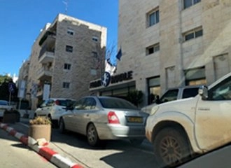 A street view of a hotel entrance, possibly in Jerusalem. The hotel is a beige stone building with the name "PARK ROYAL" visible on the facade. Several cars are parked bumper-to-bumper on the street, including a silver sedan and a white pickup truck.