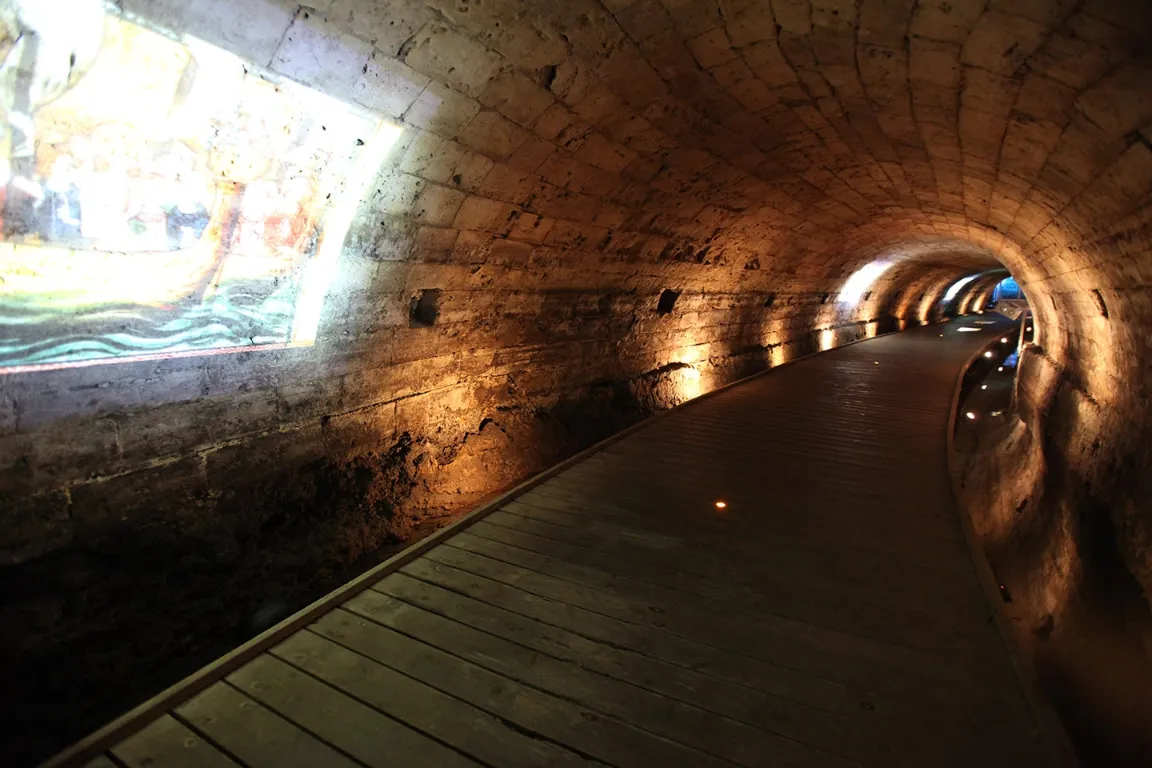 An interior view of an ancient, stone-vaulted tunnel with a wooden boardwalk. The tunnel is dimly lit by warm spotlights along the right wall and features a brightly lit cutout or projection on the left wall showing a scene.