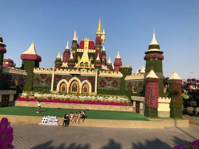 A large, elaborate castle structure in the Dubai Miracle Garden, completely covered in brightly colored flowers and green foliage. The grounds in front of the castle feature a sign that reads '#DubaiMiracleGarden' and a group of people posing for a photo.
