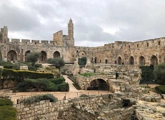 An exterior view of the Tower of David (Citadel) in Jerusalem. The photo shows the tall stone walls and ramparts, with a prominent minaret tower rising in the center. In the foreground are excavated ruins and a garden area with manicured hedges and olive trees. The sky is overcast.