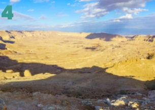 A close-up landscape of unusually bright white hills composed of soft, chalky marl and clay, sculpted by erosion into smooth, rolling mounds. The foreground is dry earth with small patches of green desert vegetation under a bright, sunny sky. This geological feature is characteristic of the Judean Desert, near the Dead Sea and Masada area.